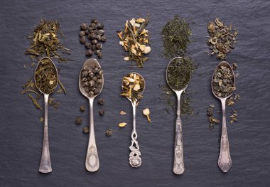 Kuru yeşil çay yaprakları gümüş kaşıklarla siyah taş arka planda.