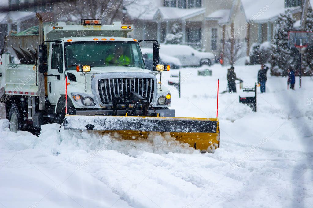 Front view of city services snowplow truck clearing roads after severe winter storm with yellow plow while children bundled for cold stand in snow in the background