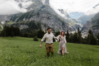 happy bride and groom holding hands and walking on alpine meadow with clouds clipart