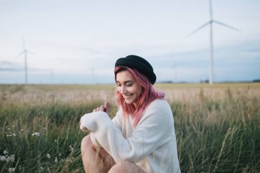 pretty girl with pink hair in white sweater and hat sitting in field with windmills clipart