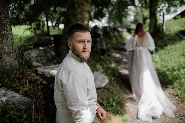 handsome young groom looking at camera and bride walking on mountain path in Alps