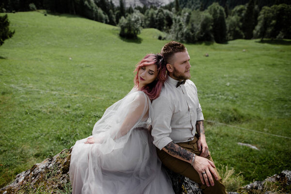 beautiful young bride and groom sitting back to back on green mountain meadow in Alps