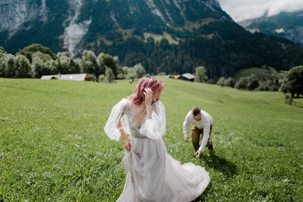 happy bride in wedding dress and groom walking on green mountain meadow in Alps