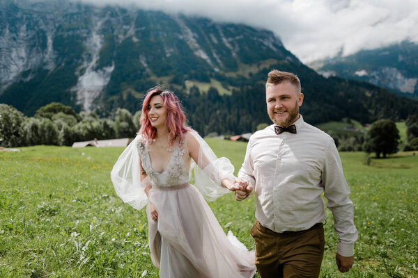 happy bride in wedding dress and groom holding hands and walking on green mountain meadow in Alps