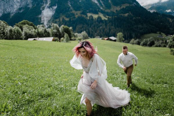 young bride with pink hair and groom walking on green mountain meadow in Alps