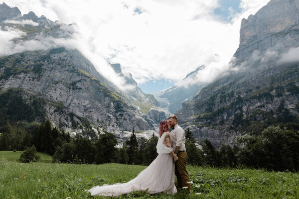 happy bride in wedding dress and groom on green mountain meadow with clouds in Alps