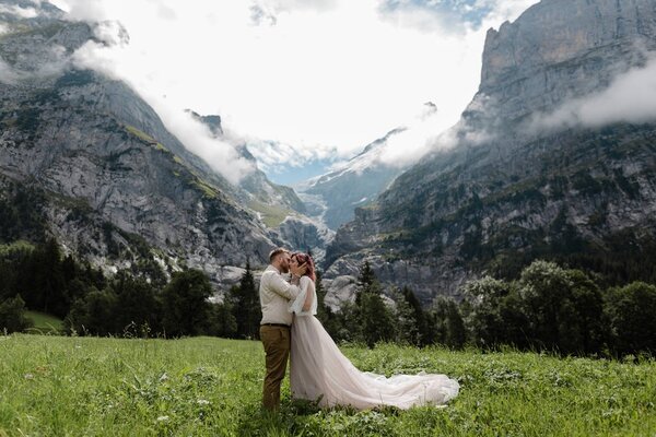 happy bride and groom hugging and kissing on green mountain meadow with clouds in Alps