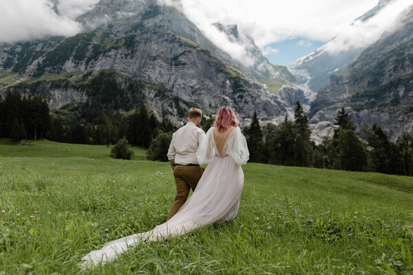 rear view of bride in wedding dress and groom holding hands and walking on green mountain meadow in Alps