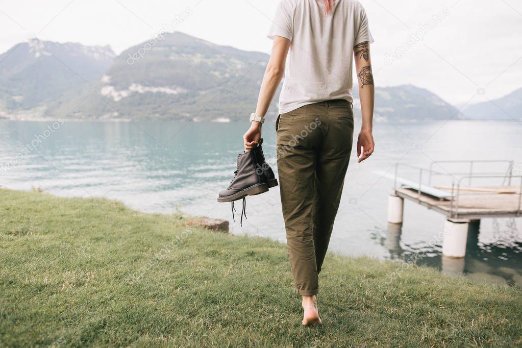 cropped shot of girl holding shoes and walking barefoot on grass near beautiful mountain lake
