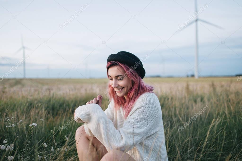 pretty girl with pink hair in white sweater and hat sitting in field with windmills