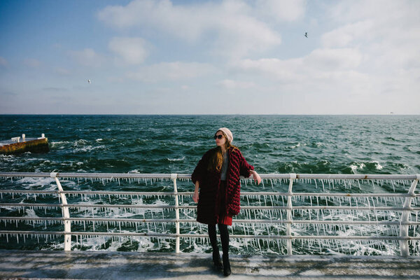 stylish girl in burgundy merino wool cardigan posing on quay near sea at winter