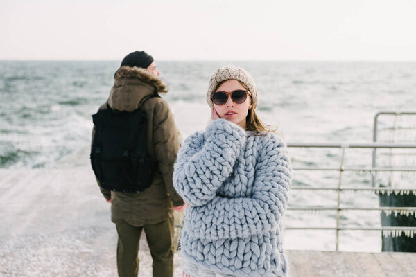 attractive stylish girl in sunglasses and merino wool sweater on winter quay, boyfriend standing behind and looking at sea