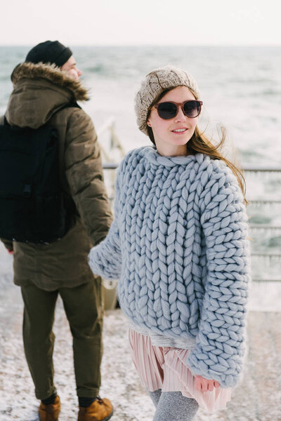 stylish girl in merino wool sweater holding hands with boyfriend on winter quay at the sea