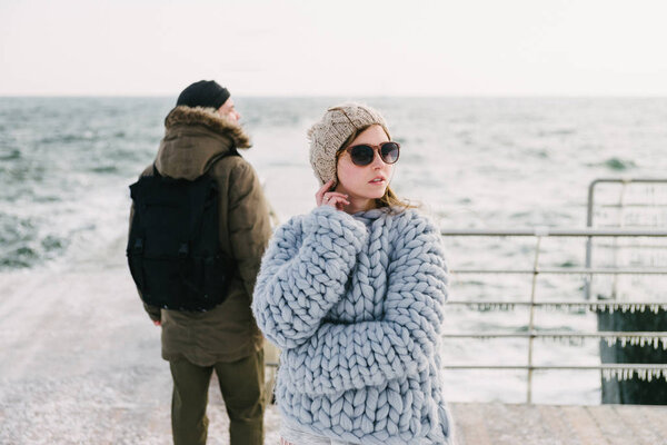 stylish girl in merino sweater on winter quay, boyfriend standing behind and looking at sea