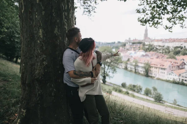 Pareja joven enamorada abrazándose cerca de un gran árbol y mirando el paisaje urbano de Berna - foto de stock