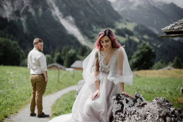 Beautiful happy young bride looking down while handsome groom standing behind in alpine valley — Stock Photo