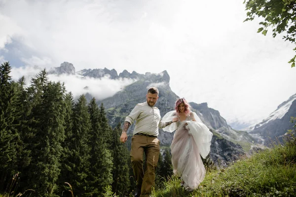 Beautiful young bride and groom holding hands and walking on green mountain meadow in  Alps — Stock Photo