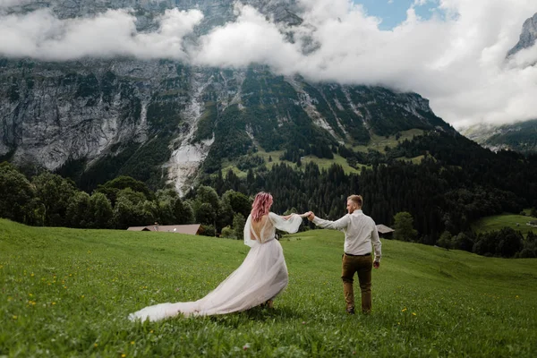 Jeune mariée et marié tenant la main sur la prairie de montagne verte avec des nuages dans les Alpes — Photo de stock