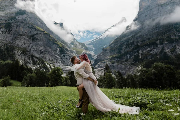 Счастливая невеста в свадебном платье и жених, обнимающий на величественном лугу с горами и облаками в Альпах — стоковое фото