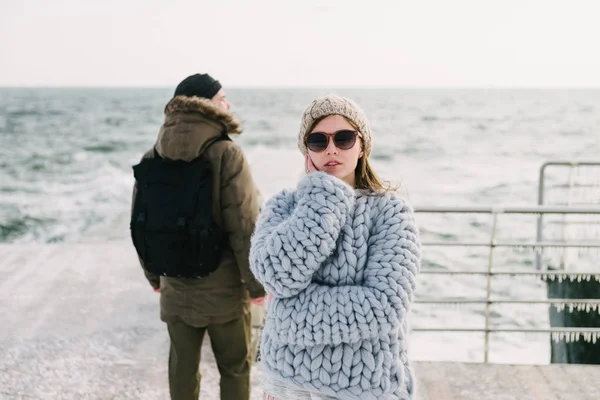 Attractive stylish girl in sunglasses and merino wool sweater on winter quay, boyfriend standing behind and looking at sea — Stock Photo