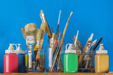 Cam kavanozdaki boya fırçaları ve plastik kaplarda akrilik boya. Mavi arka planda sanat ekipmanları