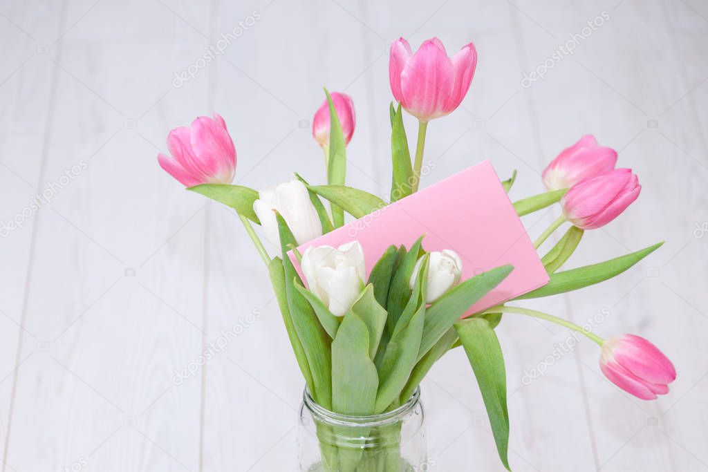 Pink tulips bouquet in glass vase on white wooden background with card. Spring background, holiday, birthday concept.