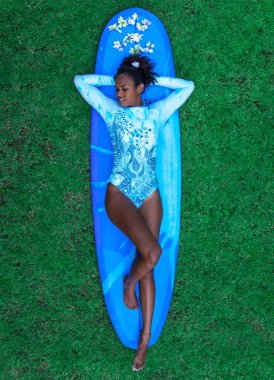 Sörfçü kız mavi tek parça bikini yeşil çimenlerin üzerinde mavi longboard arkasında yatan afro saç modeli ile