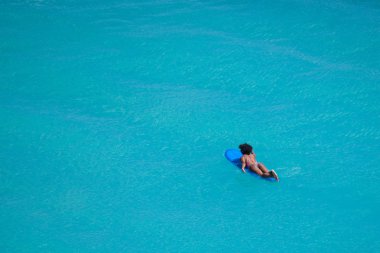 Surfer girl with afro hairstyle paddling out on surfboard in crystal clear turquoise water at Uluwatu beach, Bali, Indonesia clipart