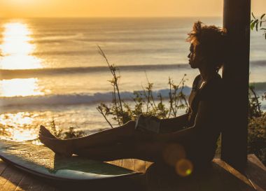 Beautiful surfer girl with afro hair style reading book on top of the cliff at sunset on the beach in front of seascape in Bali, Indonesia. copy space clipart