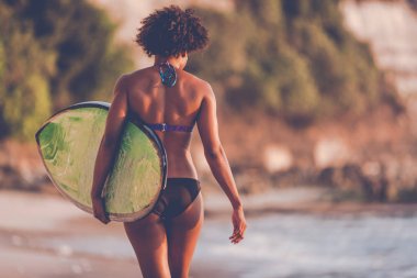 Surfer girl with afro hairstyle walking with surfboard on the Uluwatu beach at sunset in Bali, Indonesia clipart