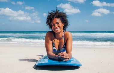 Surfer girl with afro hairstyle smiling looking at camera, lying down on blue surfboard on the white sand at Dreamland beach, Bali, Indonesia clipart