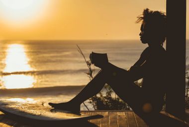 Bali, Endonezya deniz manzarası önünde sahilde gün batımında uçurumun üstünde kitap okuma afro saç stili ile güzel sörfçü kız. kopya alanı