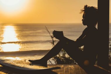 Beautiful surfer girl with afro hair style reading book on top of the cliff at sunset on the beach in front of seascape in Bali, Indonesia. copy space clipart
