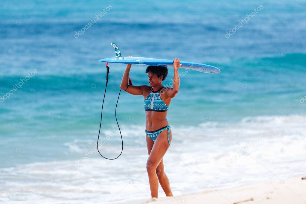 Pacific islander melanesian papuan surfer girl with afro hairstyle carrying blue longboard on head walking in the sand at Padang Padang beach, Bali, Indonesia