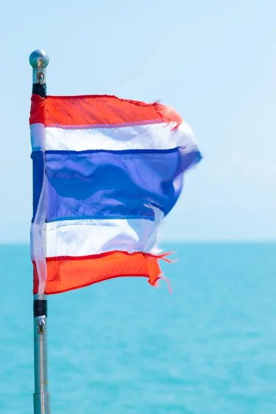 Fluttering in the wind torn Thailand flag in front of the light blue sea and sky background. Flag rod is in camera focus and the end of the flag is in motion blur