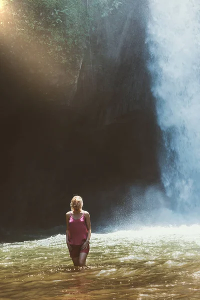 Young blonde woman with pink blouse and shorts walking in brown water river and illuminated by the sunbeam in front of the running blue waterfall in Bali island