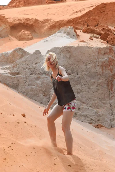 Blonde young woman with brown blouse and colorful shorts walking on red sand hills and cliffs in Mui Ne, Vietnam. Main subject is in motion blur
