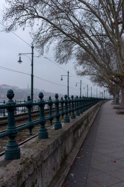 Nehir manzaralı perspektifte çıplak ağaçlar ve demiryolları ile boş sokak. Ulaşım ve seyahat konsepti. Avrupa lanscape ve şehir manzarası. Yolculuk için soğuk bir gün. Raylar, ağaçlar ve nehir boyunca yol.