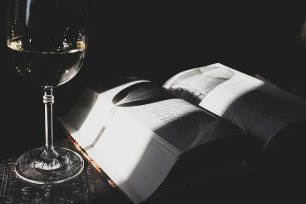 Gün ışığında filtrelenmiş açık kitap ve bir bardak beyaz şarap. Masa ahenkli görüntü üzerine kitap ve cam. Edebiyat konsepti. Evde eğitim kavramı. Okuma ve çalışma konsepti. Gün ışığında açık kitap ve bir kadeh şampanya..