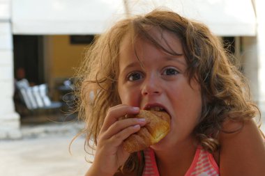 Büyük gözlü güzel küçük kız kahvaltıda kruvasan yiyor.