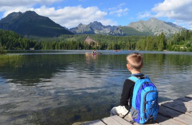 Sırtında bir sırt çantası ile bir genç çocuk hiker dağlar peyzaj (Strbske Pleso göl, yükseklik Tatras dağlar, Slovakya, Europe seyir göl kenarında oturur)
