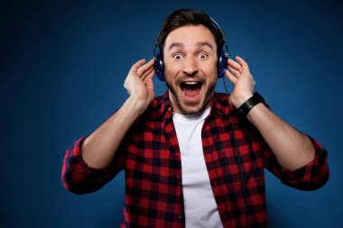 Onun telefonunu kulaklıktan müzik dinleme ve adam bir smartphone ve hava gitar çalmak iyi müziği seviyor sevinçle, favori grubu ile birlikte şarkı koyu mavi zemin üzerine izole kırmızı tişörtlü yakışıklı sakallı adam, mutlu olduğunu