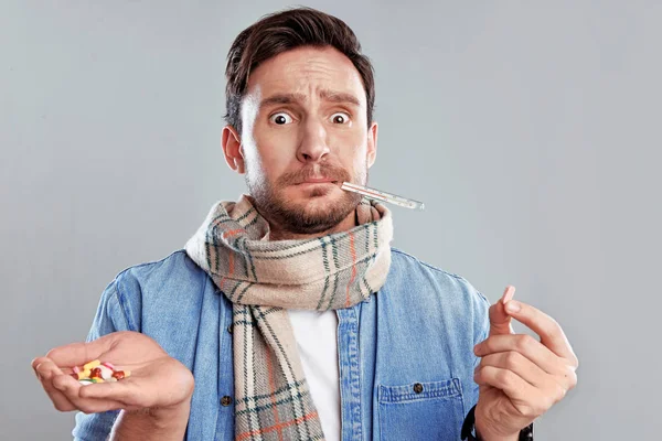 Handsome bearded man in a scarf holding thermometer and a hand full of pills isolated on a white background. Sad concerned guy in a scarf is ill and looks at the thermometer, he has a temperature