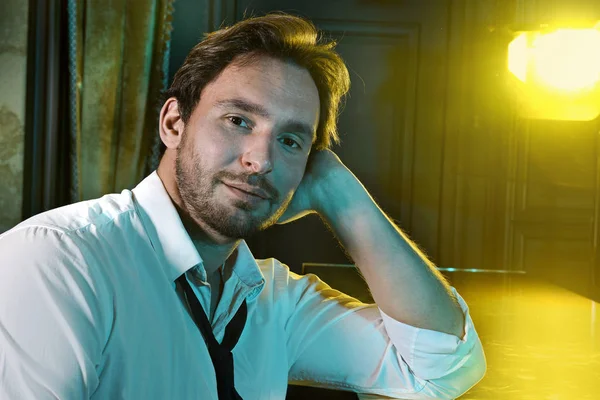 Young and handsome man sitting alone at the bar counter with a glass and a bottle. Alcoholic in the bar with a bottles around him. Beautiful Barman or waiter with a sexy look