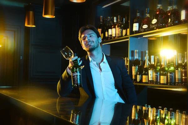 年轻英俊的男人独自坐在酒吧柜台 杯子和瓶子 酒在酒吧里 周围有个瓶子 漂亮的酒保与性感的外观 — 图库照片