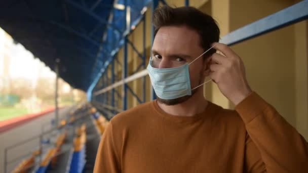 空のスタジアムに立って外科マスクの男 男は指を口に向けて咳をしながら顔の上にマスクを置く 流行性ウイルス病から守るためにマスクをしたコロナウイルスを恐れています — ストック動画