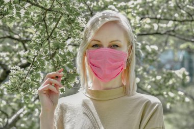 Umudu olan genç sarışın kadın, ilkbaharda çiçekli güzel bir ağacın yanında salgın ve koronavirüs salgını için karantina altındaki parkta yürüyor ve temiz hava soluyor.