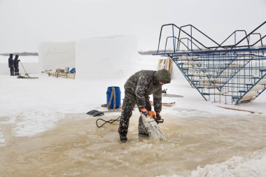 Worker in a green jacket with a hood with a chainsaw in his hands