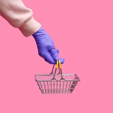 Tek kullanımlık parlak eldivenli bir kadının elinde pembe arka planda süpermarketten alınmış metal bir sepet var. Koronavirüs salgını sırasında müşterilerin güvenliği..
