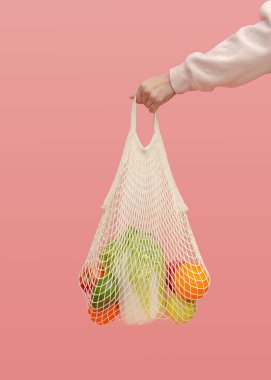 Bir kadının eli pembe arka planda sebze ve meyve dolu bir ağ torbasını tutuyor. Sıfır atık ve sağlıklı yiyecek kavramı. Naylon poşetler yerine süpermarkete gitmek için yeniden kullanılabilir bir çanta..